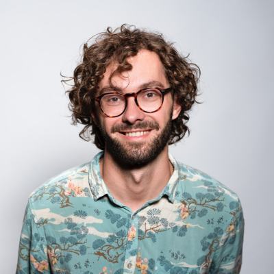 Friendly young man with curly hair and glasses smiling, wearing a floral shirt.
